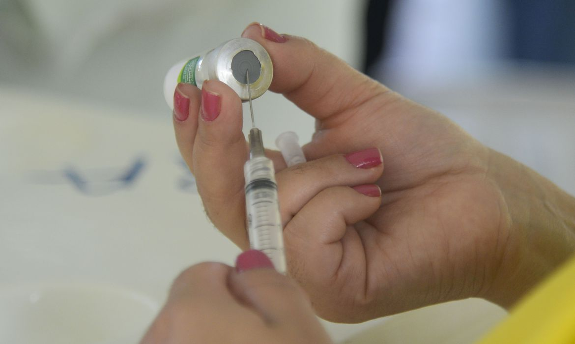 Mãos femininas enfiando agulha de uma seringa em um frasco de vacina.