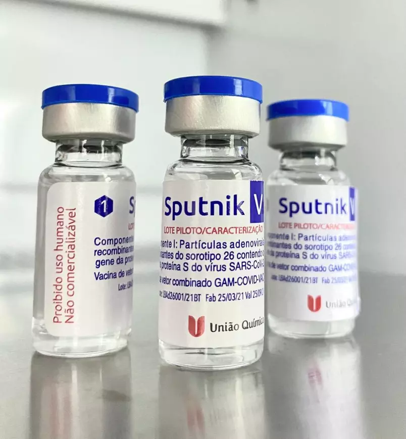 Imagem de três frascos de vacinas Sputinik, sobre uma mesa, com rótulo branco e texto azul marinho e vermelho, além de lacre metálico com topo de plástico azul.