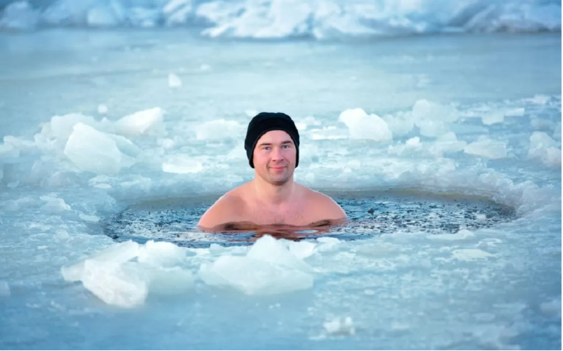 Homem apenas de touca nadando em um lago congelado. A imagem mosta ele submerso em água, somente com os ombros e a cabeça de fora d'água.