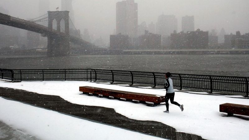 Foto de um local nevando, à margem de um rio. Uma pessoa faz exercícios com roupas de frio.