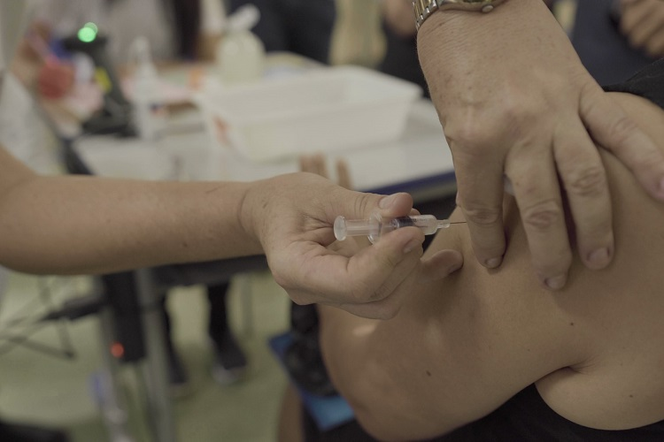 Foto de uma pessoa sendo vacinada. Ela está sentada e a imagem foca em seu braço esquerdo, enquanto um profissional da enfermagem enfia a agulha da seringa em seu braço.