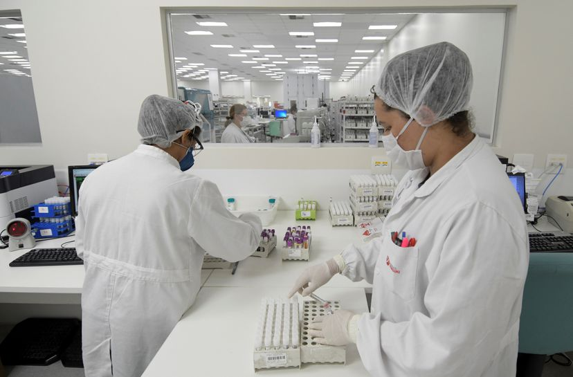 A imagem mostra duas funcionárias vestindo equipamentos de proteção individual, organizando tubos de exames para covid-19 em uma sala.